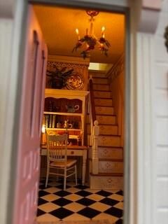 Narrow hallway with black-and-white checkered floor, wooden chair, shelving, staircase, and warm amber lighting