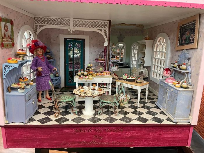 Miniature bakery display with pink walls, checkered floor, and a doll arranging desserts on tables