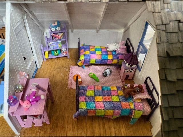 Miniature dollhouse bedroom with two colorful beds, a pink desk, and toy shelves on a wooden floor