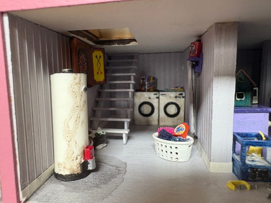 Small laundry room with washer, dryer, staircase, water heater, and laundry basket in a cluttered basement.