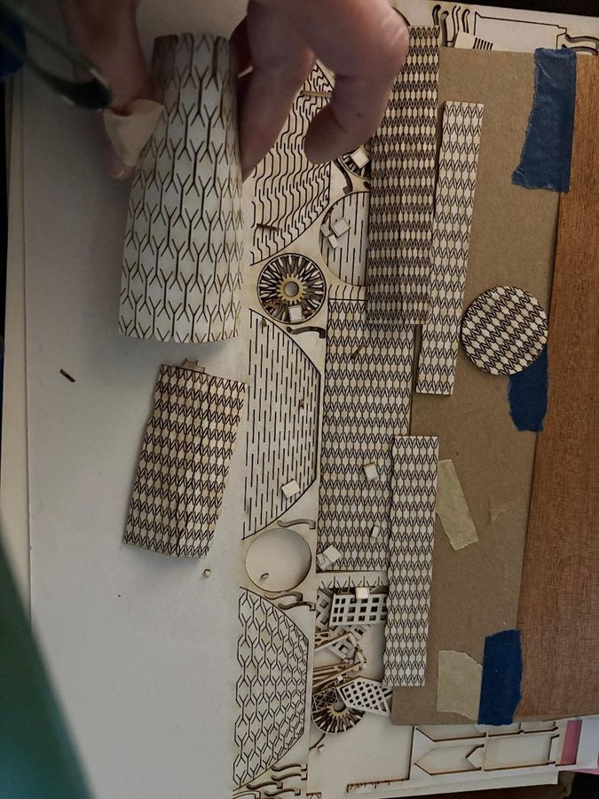 Hand placing patterned paper cutouts on a brown board with blue tape and fabric swatches