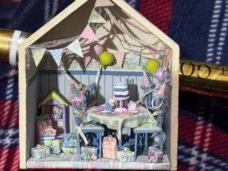 Miniature garden-themed dollhouse with pastel furniture, bunting, and tiny balloons on a shelf