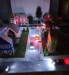 Miniature village street at night with small houses and cars lit by red and white lights