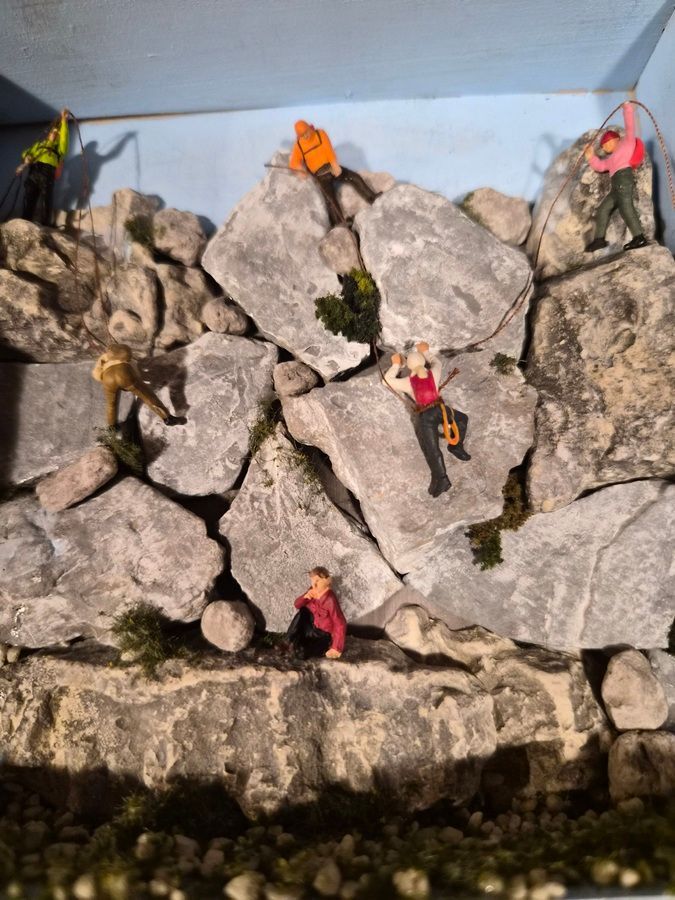 Tiny climbers on gray rocks in a terrarium, with one orange figure at the top and a small plant at left.
