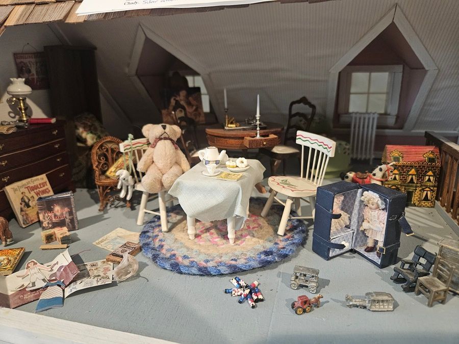 Miniature dollhouse attic room with table, chairs, toys, books, and a fireplace display