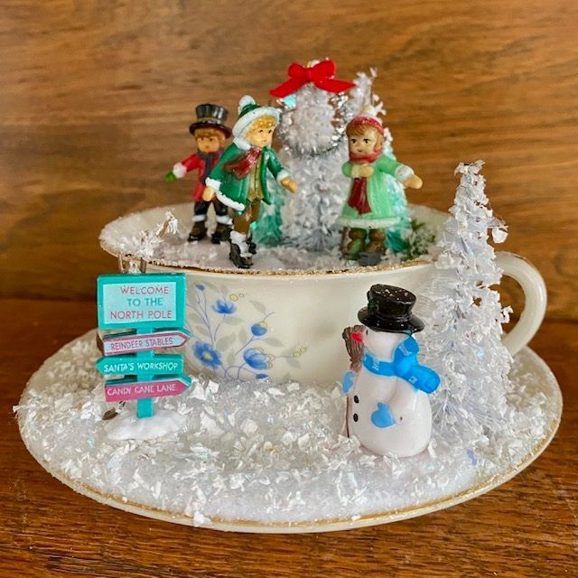 Snowy holiday teacup display with a snowman, elf figurines, and a tiny festive sign on a saucer