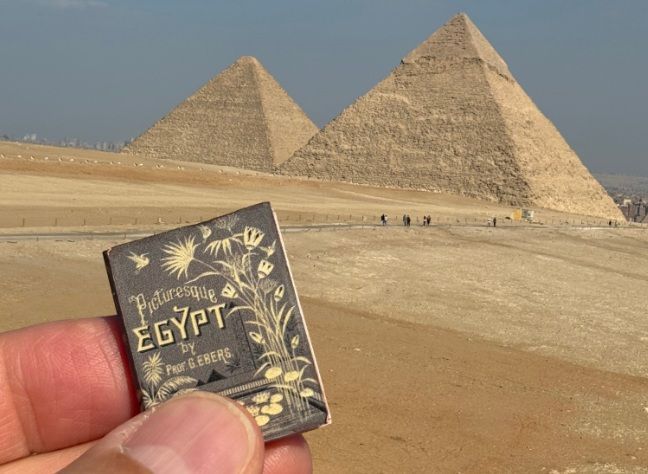 Hand holding a small “Egypt” guidebook in front of the pyramids at Giza.