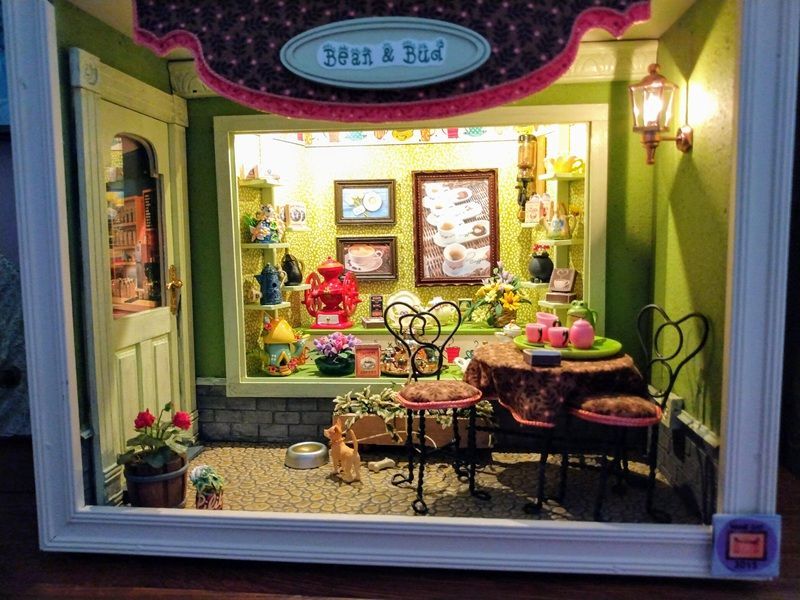 Tiny colorful shop display with mini tables, chairs, and framed art in a bright green storefront window
