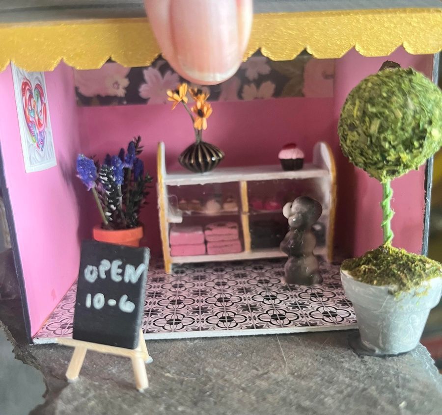 Miniature pink shop display with open sign, shelf, potted tree, and patterned floor inside a small diorama.