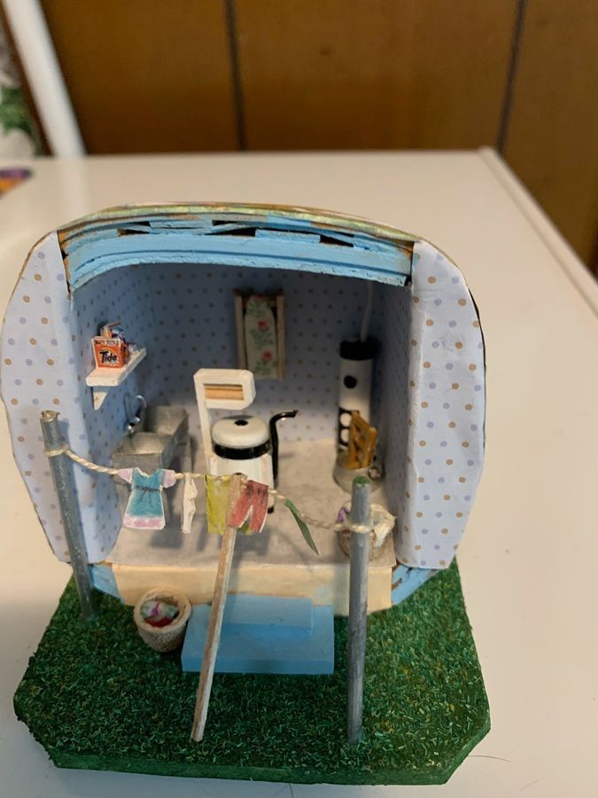 Miniature bathroom dollhouse with shower, toilet, sink, and hanging clothes on a green base.