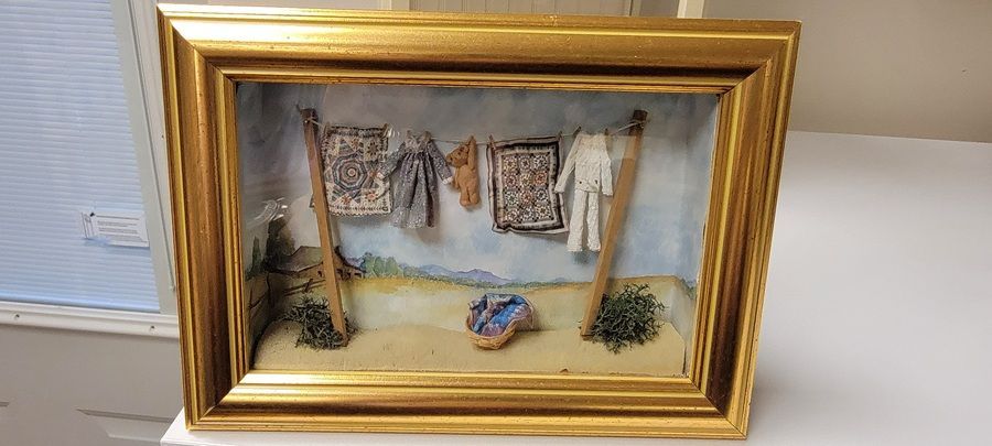 Framed miniature textile scene with hanging laundry and a small blue bundle on a sandy floor
