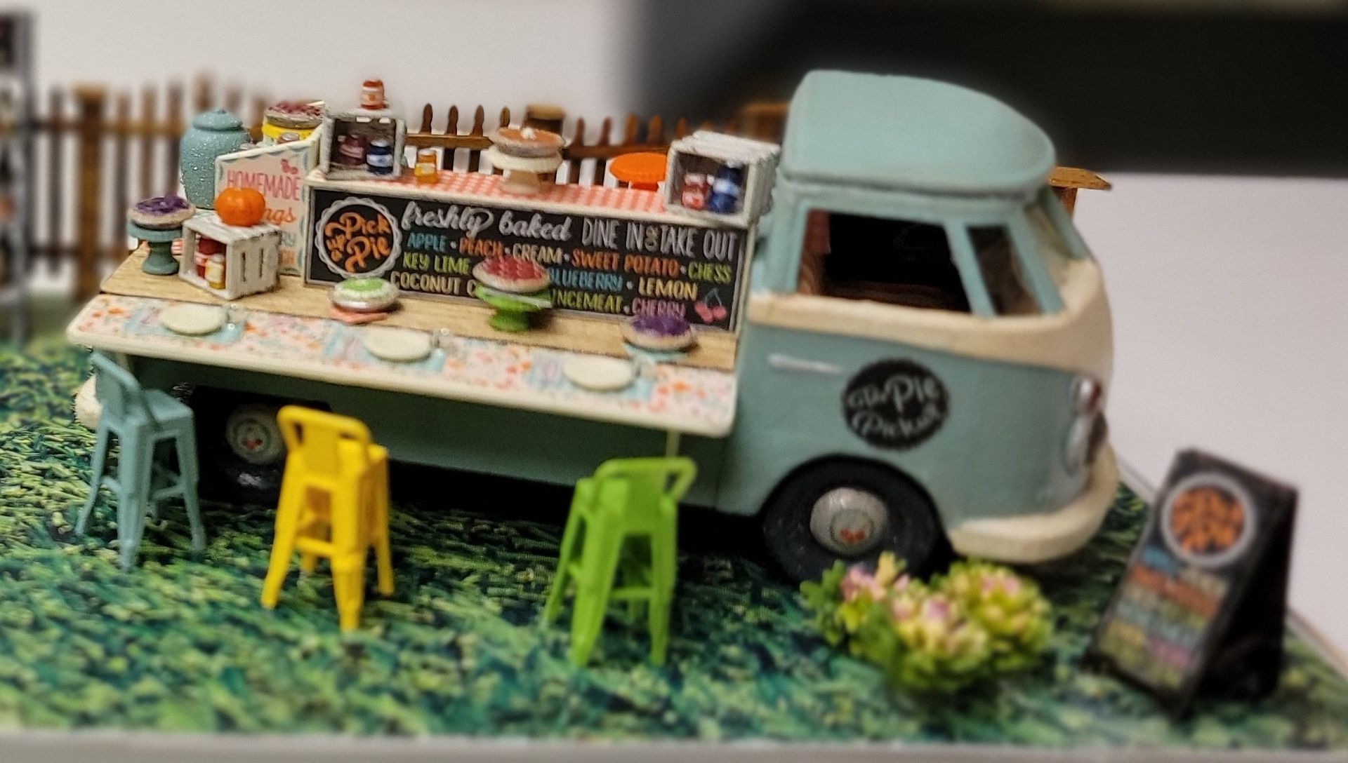 Miniature food truck scene with colorful menu board, stools, and a small drinks display on a grassy base