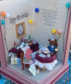 Miniature birthday-themed room display with tables, balloons, gifts, and a framed portrait on a pink base
