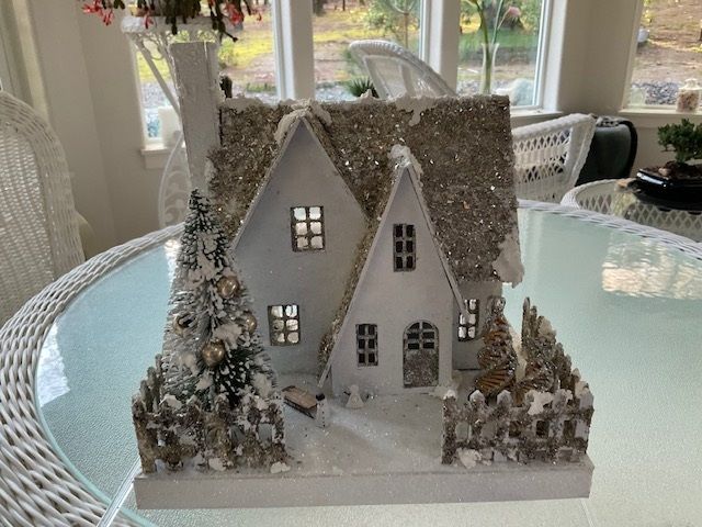 Snow-covered miniature house with lit windows on a glass table by a bright window