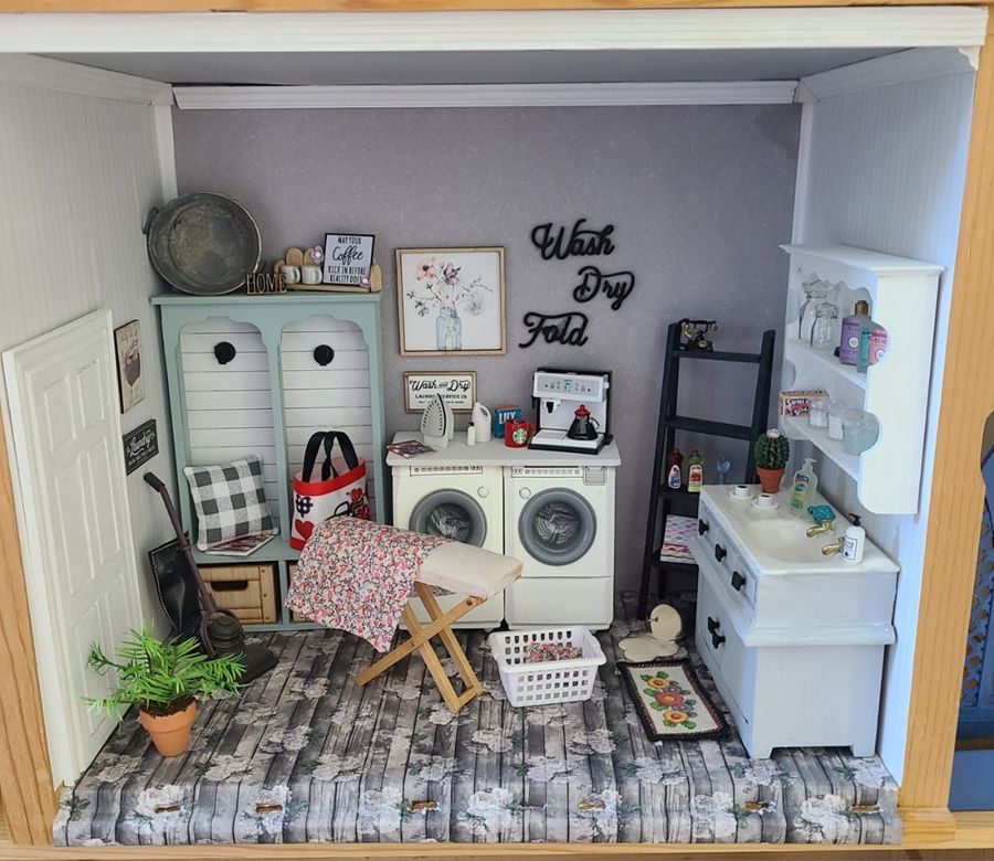 Miniature laundry room with washer, dryer, ironing board, shelves, plants, and wall decor in a dollhouse.