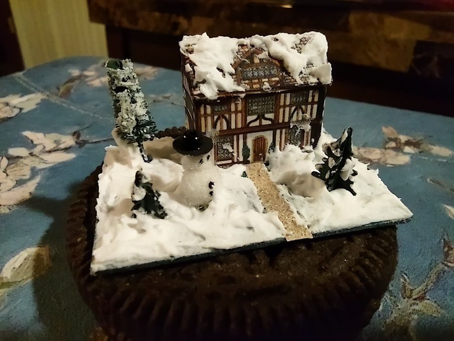 Snow-covered miniature house on a dark base, with tiny trees and a path on a tablecloth.