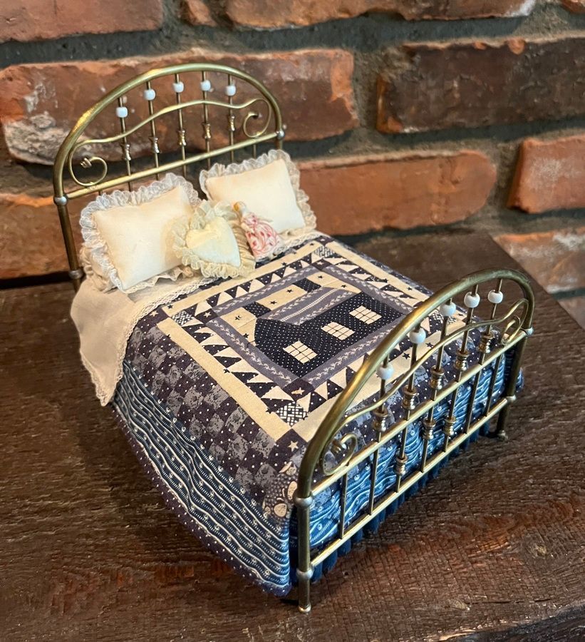 Miniature bed with blue-and-cream quilt and pillows, shown on a dark surface against a brick wall.