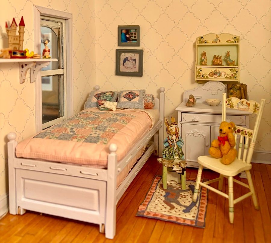 Miniature bedroom with a white bed, tiny desk, chair with teddy bear, and shelves on patterned wallpaper.