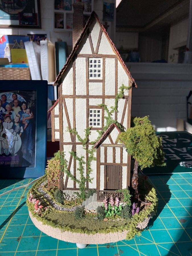 Tall narrow model house with white walls, brown trim, green vines, and a circular garden base.