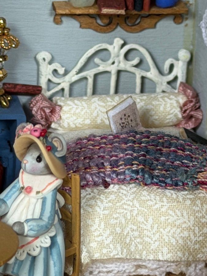 Miniature bed with white ornate headboard, cream bedding, patterned quilt, and a doll beside it