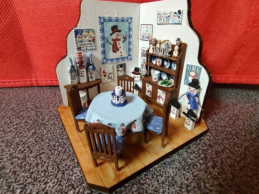 Miniature dining room with blue tablecloth, wooden chairs, shelves, and framed wall art on a wooden base.