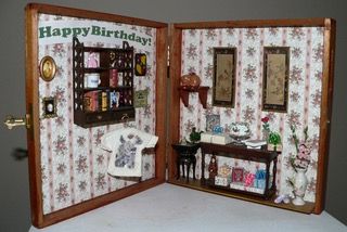 Miniature open dollhouse with floral wallpaper, framed art, shelves, and a “Happy Birthday” sign on the left wall