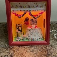 Miniature room display in a red frame with orange walls, garlands, a chair, and a table covered in a white cloth.