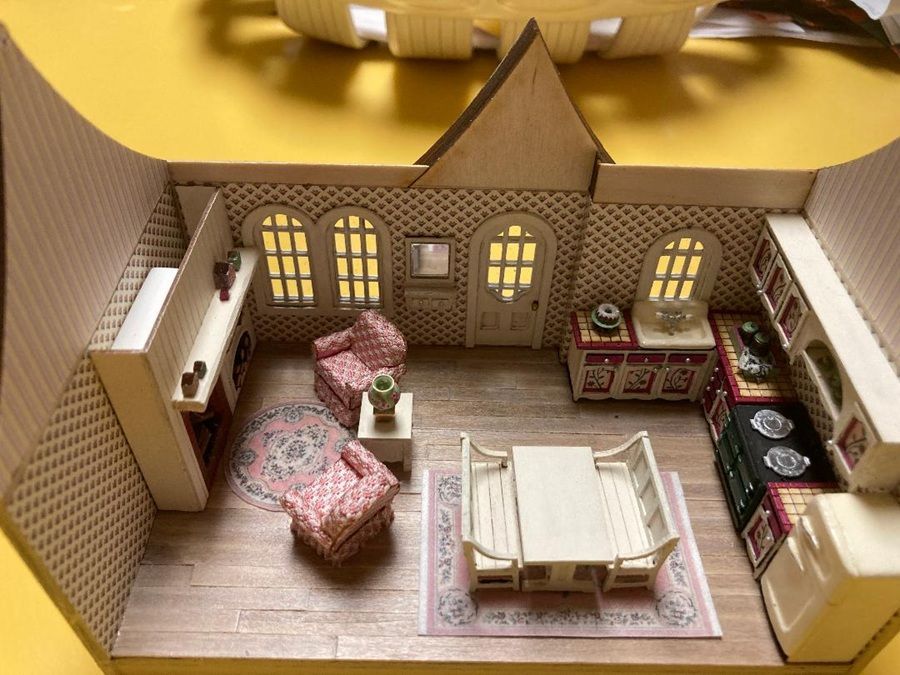 Miniature dollhouse living room with pink armchairs, dining table, cabinets, and arched windows.