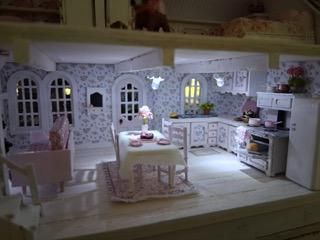 Miniature white dollhouse kitchen with dining table, cabinets, arched windows, and floral wallpaper