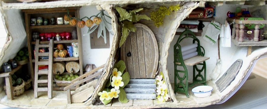 Whimsical miniature fairy house with arched wooden door, shelves, ladder, flowers, and mossy tree trunk