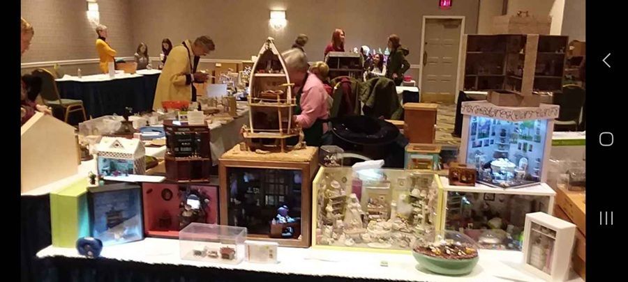 Indoor craft fair display of miniature holiday scenes and model houses on tables with people browsing in the background