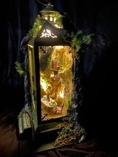 Illuminated decorative lantern with plants and a small figurine inside, against a dark background
