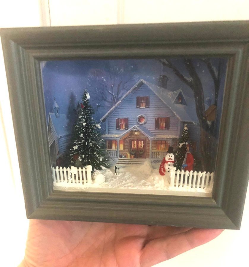 Framed miniature winter house scene with snow, lit windows, a tree, and a snowman behind a white fence