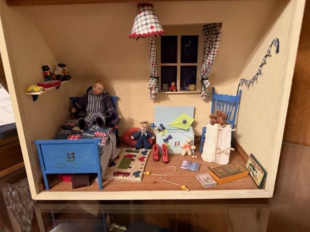Miniature dollhouse bedroom with blue bed, window, rug, and small furniture on a wooden shelf.