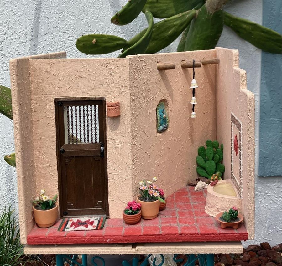 Miniature courtyard diorama with a brown door, potted plants, red tile floor, and hanging wind chime.