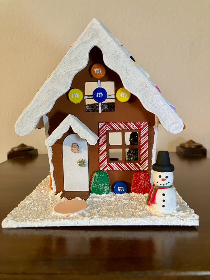 Miniature gingerbread house with white icing roof, candy decorations, and a small snowman on a base plate