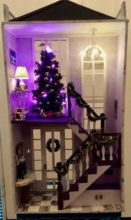 Dollhouse living room with a lit Christmas tree, purple lamp glow, and staircase with garland.