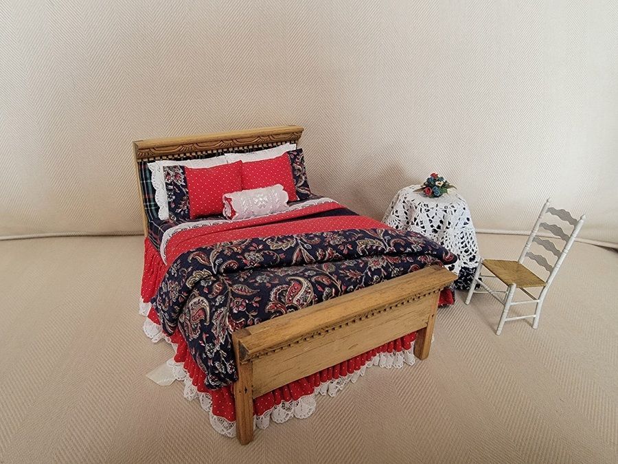 A small bed with red and floral bedding beside a white chair and a side table in a sparse room.