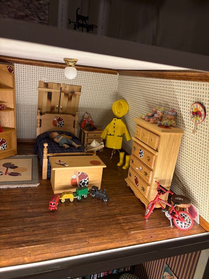 Miniature dollhouse room with wooden furniture, yellow figurine, toy table, dresser, and red bike on a brown floor.
