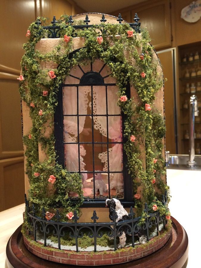 Decorative miniature storefront with pink flowers, greenery, and a tall arched window in a display case