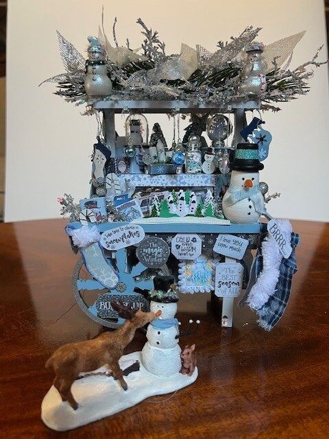 Decorated winter-themed miniature display with snowman and deer figurines on a wooden table.