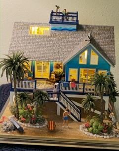 Miniature blue house diorama with lit windows, porch, stairs, palm trees, and a small figurine in front.