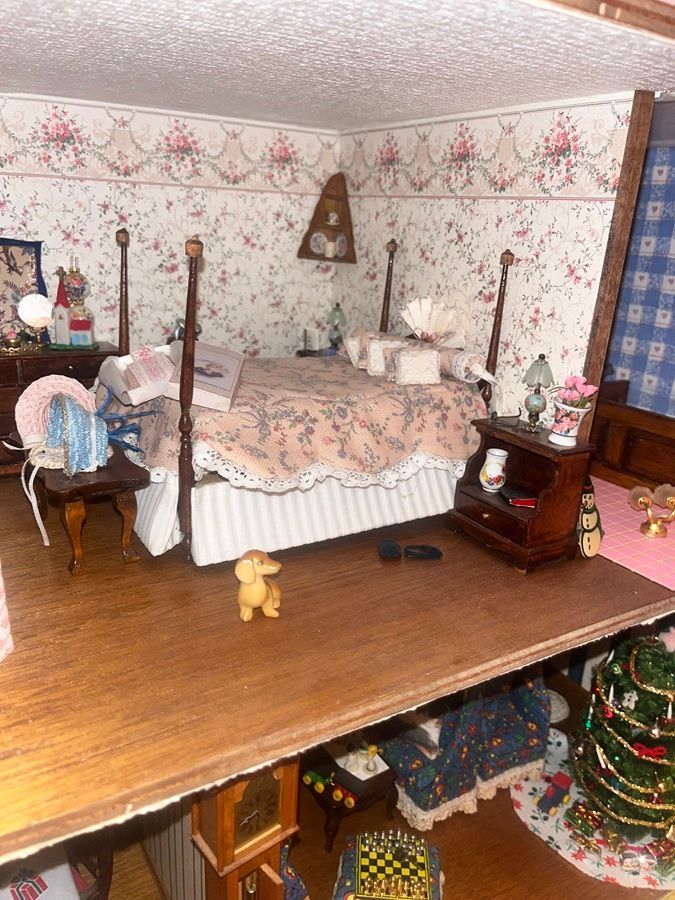 Miniature bedroom dollhouse with floral wallpaper, bed, chair, and Christmas tree on a wooden shelf.