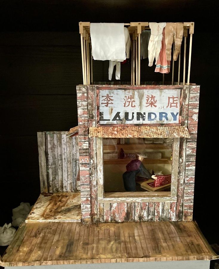 Miniature laundry shop with hanging clothes and a person inside, painted in weathered wood and brick tones