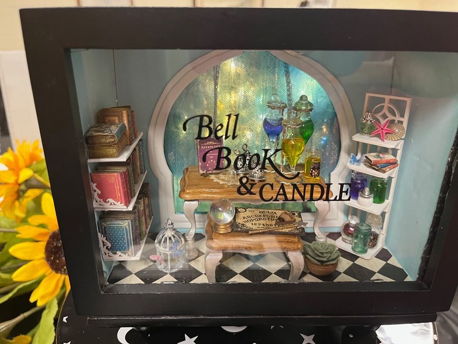 Miniature book and candle shop display in a black frame with colorful shelves and checkered floor.