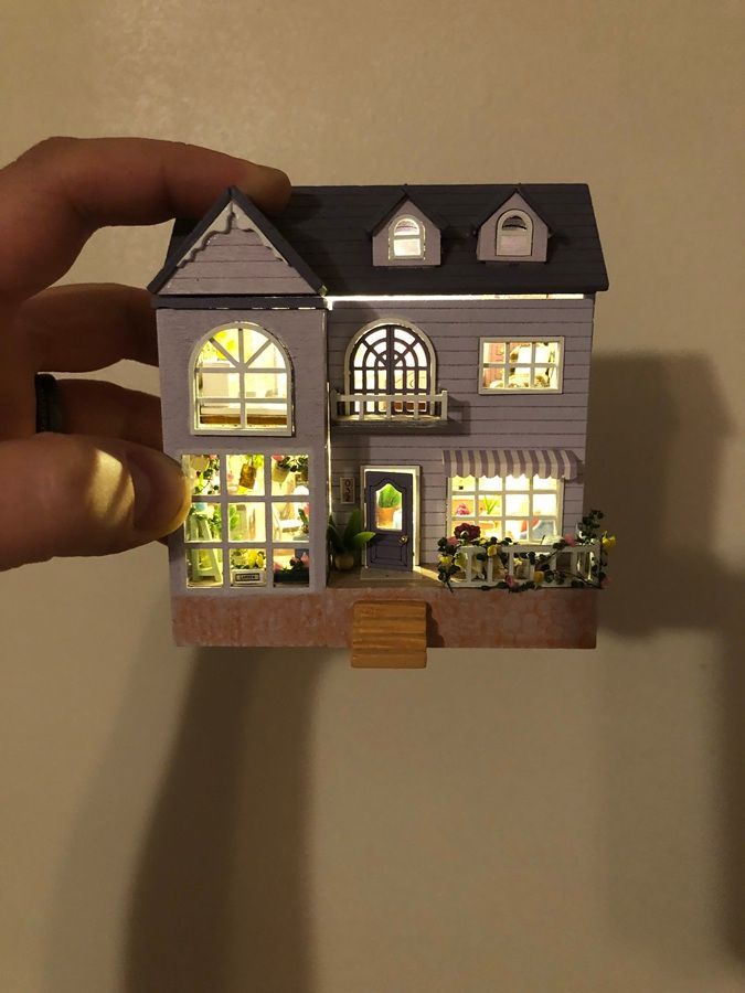Hand holding a lit miniature dollhouse with glowing windows and gray roof against a plain wall