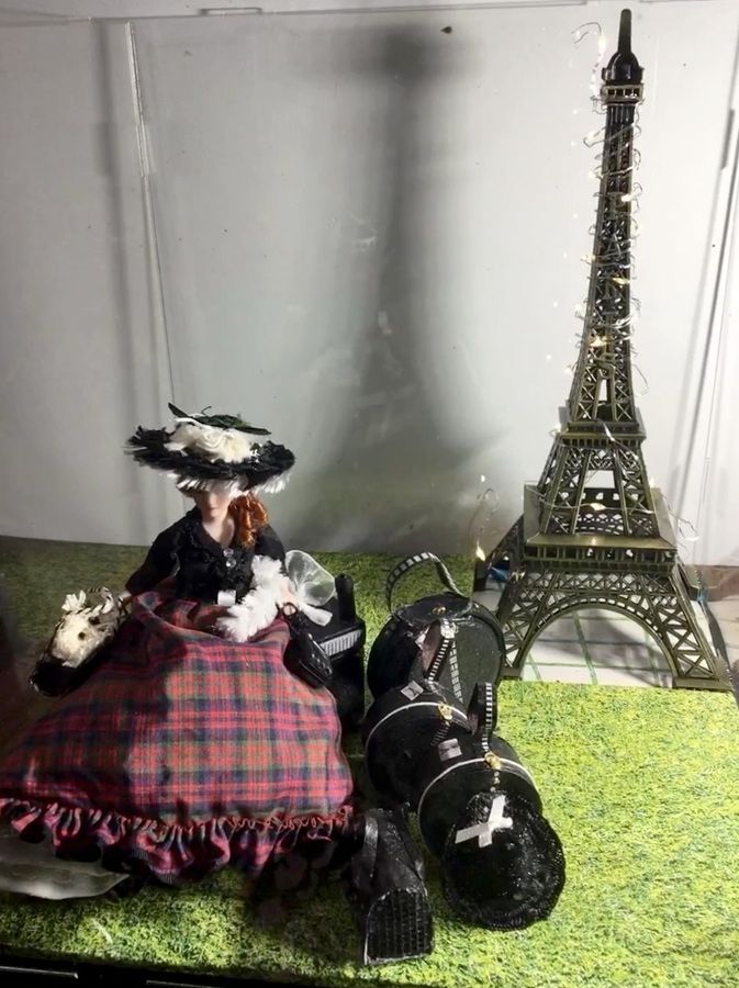 Doll in plaid dress beside black cat figurines, with a miniature Eiffel Tower on a green surface.