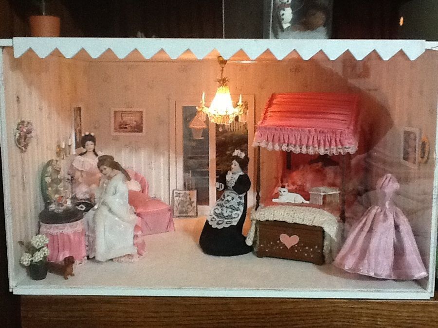 Miniature dollhouse bedroom with pink decor, dolls, chandelier, canopy bed, and lace curtains