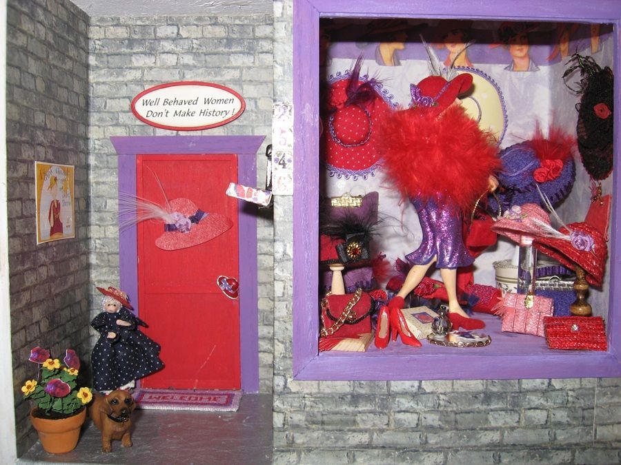 Miniature brick storefront with red door and colorful window display of mannequins and feathers