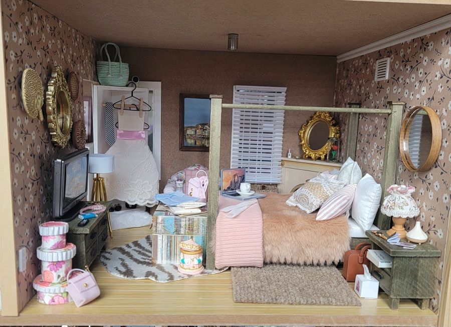 Miniature room display with pink and cream decor, beds, pillows, mirrors, and baskets in a cozy boutique setting